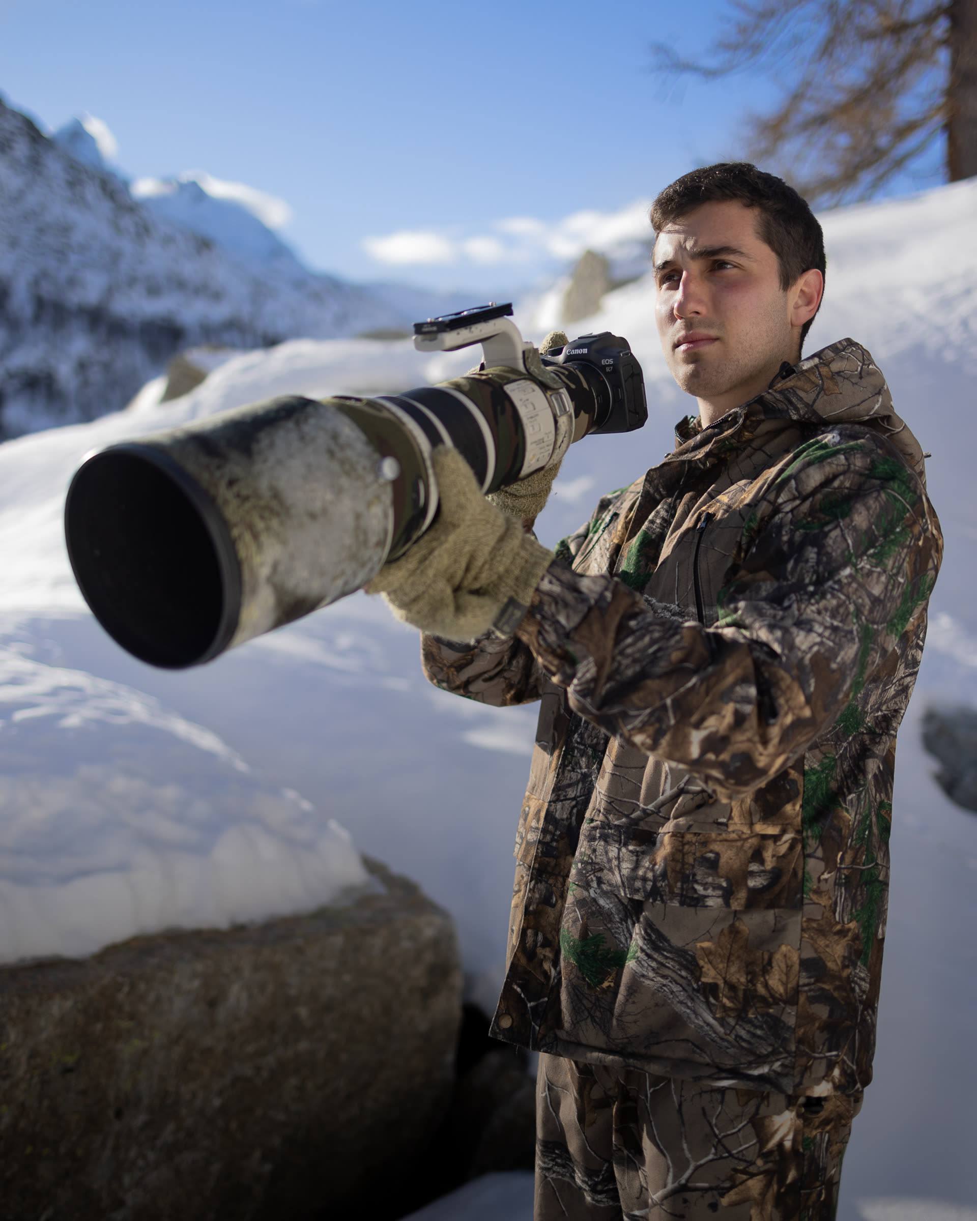 Matteo Gaglio in tuta mimetica inquadra con un teleobiettivo, tra le montagne innevate